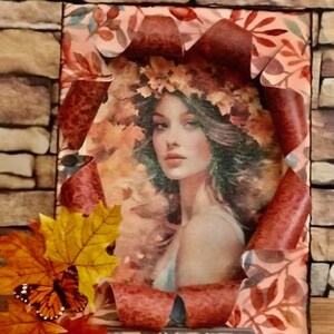 May include: A framed artwork featuring a woman with a crown of leaves, surrounded by a decorative border of autumn leaves in shades of red, orange, and brown.