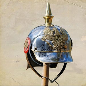 May include: A polished silver-colored helmet with a gold-colored spike and eagle emblem. The helmet has a red and gold side detail and a black chin strap. The text "Medieval Replicas" is visible on the helmet.