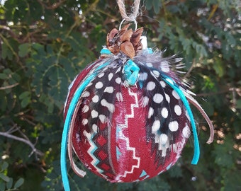 May include: A red, white, and black patterned ornament with turquoise accents and feathers. The ornament is decorated with a pine cone and a turquoise bead.