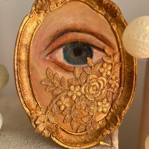 May include: Oval-shaped artwork featuring a painted eye with a blue iris, framed in gold. Gold floral lace partially covers the eye. The frame has ornate detailing and a distressed gold finish, creating a vintage aesthetic.