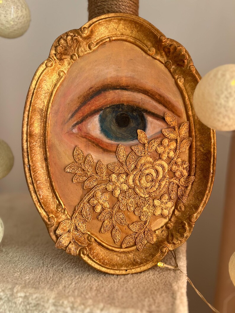 May include: Oval-shaped artwork featuring a painted eye with a blue iris, framed in gold. Gold floral lace partially covers the eye. The frame has ornate detailing and a distressed gold finish, creating a vintage aesthetic.