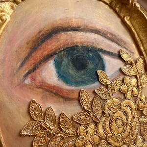 May include: A framed artwork featuring a painted eye with blue iris and detailed eyelashes. The eye is surrounded by a gold-colored, ornate frame and embellished with gold lace floral details.