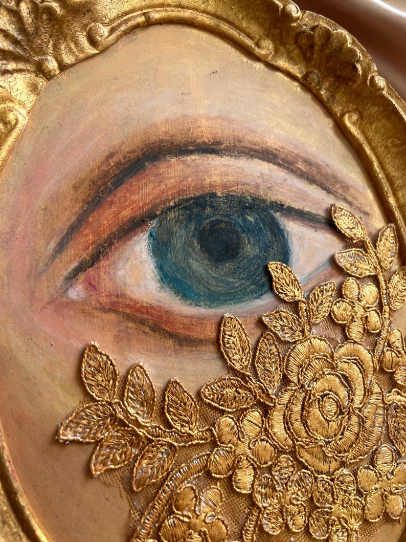 May include: A framed artwork featuring a painted eye with blue iris and detailed eyelashes. The eye is surrounded by a gold-colored, ornate frame and embellished with gold lace floral details.
