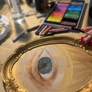 May include: A gold-framed oval painting of an eye, with a dark blue iris and peach-colored eyelids. A hand with red nail polish holds a paintbrush, and art supplies are in the background.