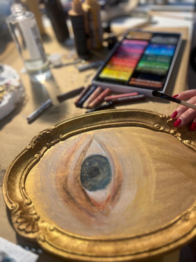 May include: A gold-framed oval painting of an eye, with a dark blue iris and peach-colored eyelids. A hand with red nail polish holds a paintbrush, and art supplies are in the background.