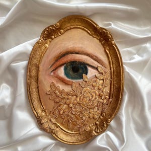 May include: An oval-shaped artwork featuring a painted eye with a blue iris, framed in gold. A gold floral embellishment partially covers the eye. The artwork is set against a white, satin-like background.