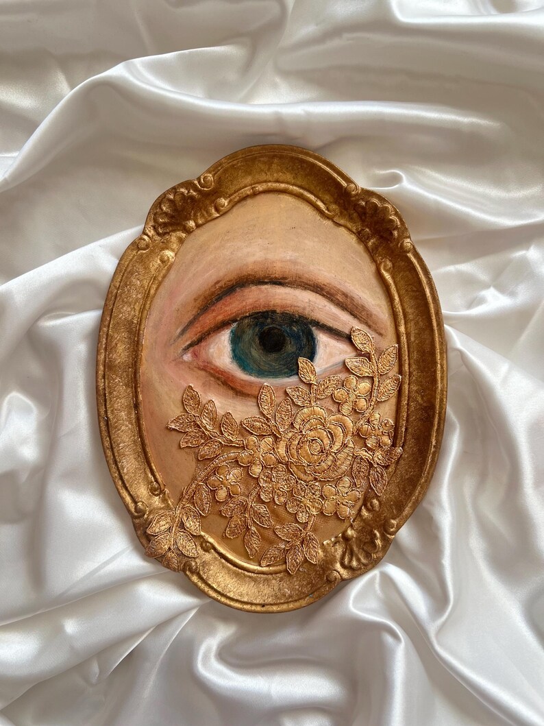 May include: An oval-shaped artwork featuring a painted eye with a blue iris, framed in gold. A gold floral embellishment partially covers the eye. The artwork is set against a white, satin-like background.