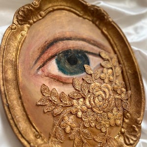 May include: Oval-shaped artwork featuring a painted eye with blue-green iris, framed in gold. A gold floral lace design partially covers the eye. The frame has an ornate, textured design, adding a vintage aesthetic.