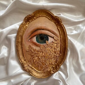 May include: A gold-framed artwork featuring a painted eye with a teal iris and a floral embellishment. The eye is set against a beige background, with the frame and floral details in a warm gold tone. The artwork is displayed on a white satin surface.
