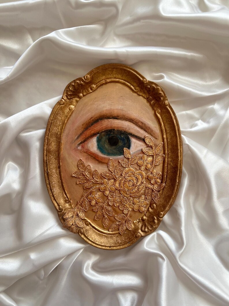 May include: A gold-framed artwork featuring a painted eye with a teal iris and a floral embellishment. The eye is set against a beige background, with the frame and floral details in a warm gold tone. The artwork is displayed on a white satin surface.