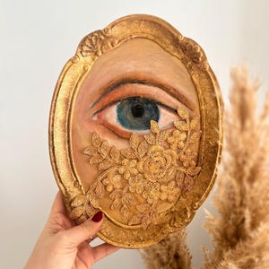 May include: Oval-shaped artwork featuring a painted eye with blue iris, framed in gold. Gold floral embellishments adorn the lower right. The artwork is held against a neutral background, showcasing the intricate details and warm color palette.