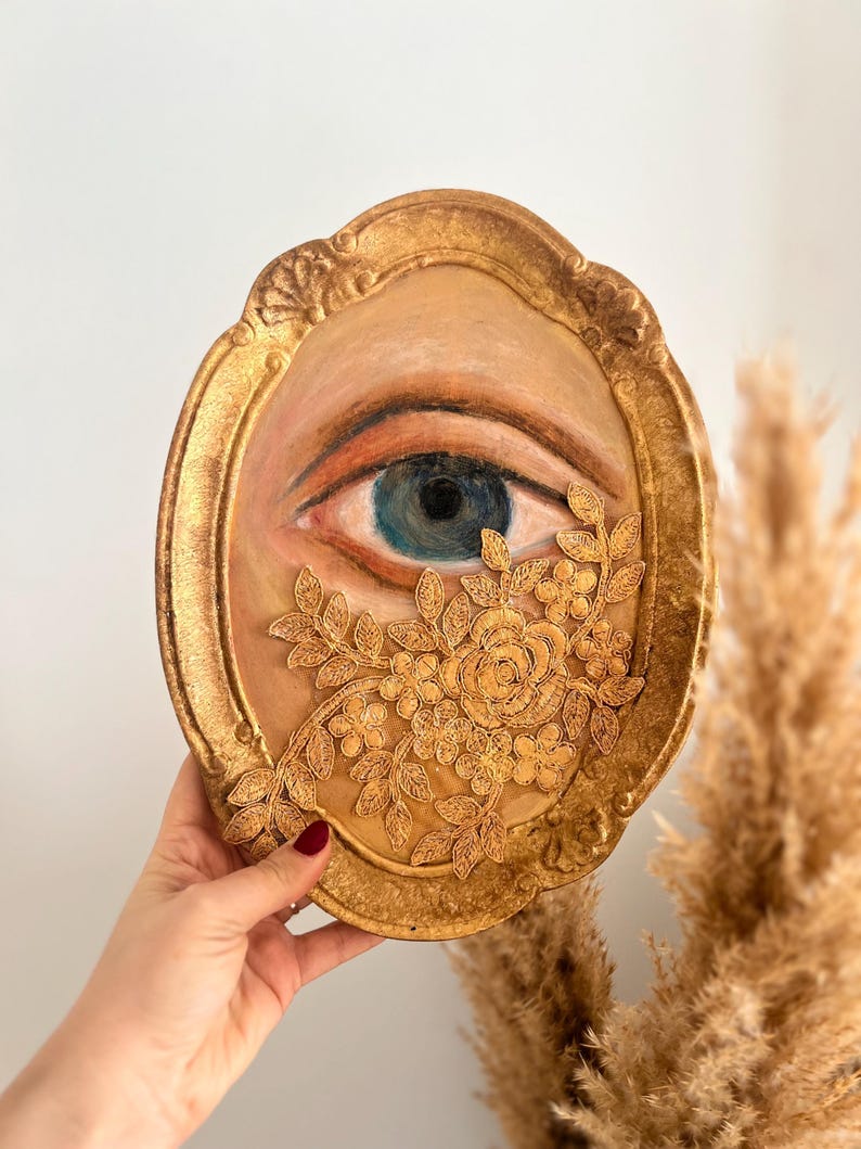 May include: Oval-shaped artwork featuring a painted eye with blue iris, framed in gold. Gold floral embellishments adorn the lower right. The artwork is held against a neutral background, showcasing the intricate details and warm color palette.