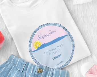 Può includere: Una maglietta bianca con una grafica circolare con la scritta "Sleeping Giant Thunder Bay Ontario CANADA" è abbinata a pantaloncini a righe blu e bianche, scarpe rosa a pois e un portachiavi con pompon rosa. Un fiocco bianco completa l'ensemble.