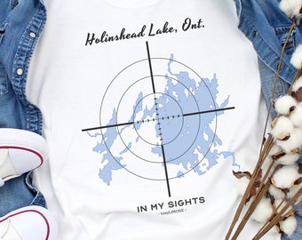Può includere: T-shirt bianco con grafica mappa blu e la scritta "Holinshead Lake, Ont. IN MY SIGHTS". La maglietta è abbinata a una giacca di jeans e scarpe da ginnastica bianche con dettagli rossi e blu. Steli di cotone disposti su una superficie di legno chiaro.
