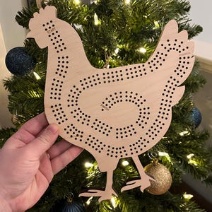 Op de afbeelding: Een houten cribbagebord in de vorm van een kip met de woorden "START" en "FINISH". Het bord is lichtbruin met talrijke kleine gaatjes voor de pinnen. Het hangt aan een kerstboom versierd met blauwe en gouden ornamenten.