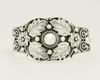 May include: Silver-toned cuff bracelet with floral and leaf design. The bracelet features detailed leaves, flowers, and circular accents. The central design element is a circular shape with a pearl-like center.