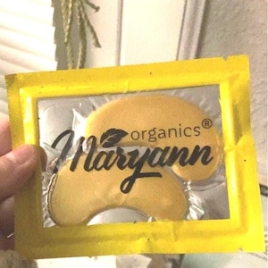 May include: A yellow pouch with a clear window showing two gold under-eye patches. The pouch has the text "organics® Maryann" printed on it.