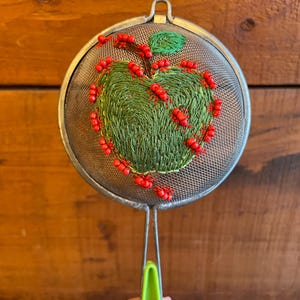 以下が含まれることがあります： 緑と赤のリンゴの刺繍デザインが施された、丸い銀色の茶こし。リンゴは緑色の糸と赤いビーズで刺繍されています。ハンドルには緑色のプラスチックグリップが付いています。