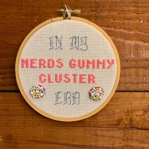 May include: A cross-stitch hoop with the words "IN MY NERDS GUNNY CLUSTER ERA" in pink and blue thread. Two clusters of colorful beads are attached to the fabric. The hoop is set against a wooden background.