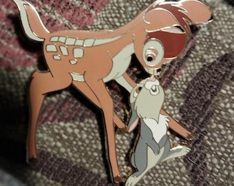 Peut inclure: Une épingle dorée représentant Bambi le cerf et Panpan le lapin du film Bambi de Disney. Bambi est brun avec des taches blanches et se penche pour embrasser Panpan, qui est gris avec un ventre blanc et regarde Bambi.