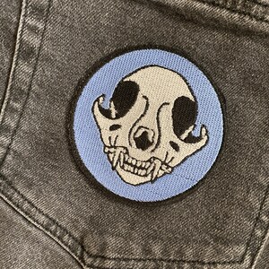 Op de afbeelding: Een blauwe en witte geborduurde patch van een katten schedel met een tanden glimlach. De patch is genaaid op een donkergrijze denim zak.