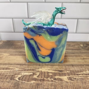 May include: A handmade soap bar with a fantasy theme. The soap features a green and blue dragon figurine on top. The soap itself has a marbled design with blue, green, and orange colors. The soap is sitting on a wooden surface.