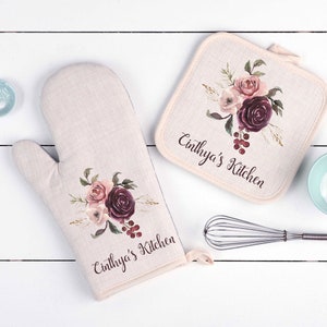 May include: A set of two beige kitchen pot holders with a floral design and the text "Cinthya's Kitchen".