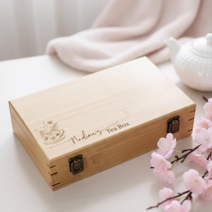 May include: A rectangular bamboo tea box with a decorative tea cup and floral design. The box is labelled "Nadine's Tea Box" in script. The box has metal clasps and hinges. Pink flowers and a white teapot are in the background.