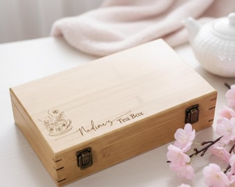 May include: A rectangular bamboo tea box with a decorative tea cup and floral design. The box is labeled "Nadine's Tea Box" in script. The box has metal clasps and hinges. Pink flowers and a white teapot are in the background.