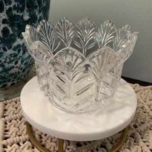 May include: A clear, cut-glass bowl with a leaf design, resting on a white marble-topped stand with a gold-colored metal base. The bowl's edges have a scalloped shape. The stand sits on a beige macrame placemat.