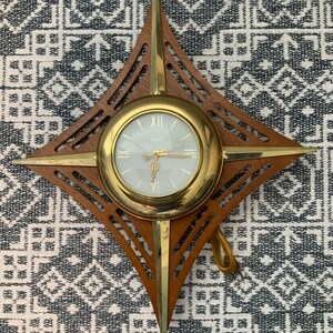 May include: A gold and brown starburst-shaped wall clock with a white face and black hands. The clock has a wood frame and a gold metal rim around the face.