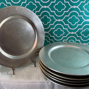 May include: A set of silver-colored charger plates. One plate is displayed upright on a decorative stand, while a stack of five plates rests on a white tablecloth. The plates have a metallic sheen and a simple, elegant design.