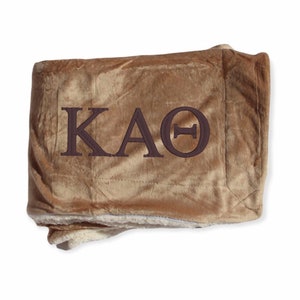 Op de afbeelding: Een bruin dekbed met de geborduurde letters KAOΘ in donkerbruin. De deken heeft een zachte, pluche textuur en een lichtere onderkant. De deken is opgevouwen en lijkt van een warm materiaal te zijn.