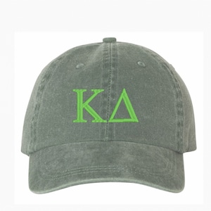 May include: A sage green baseball cap featuring the embroidered letters "K" and a Greek delta symbol in bright green. The cap has a curved peak and a fabric strap closure. The hat is made of a soft, washed cotton material.