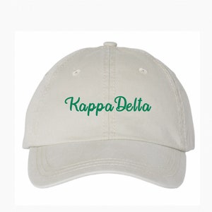May include: A light beige baseball cap featuring the green script text "Kappa Delta" on the front. The cap has a curved peak and a button on top. The hat is made of a soft fabric.