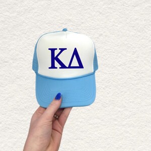 May include: A light blue and white trucker hat featuring the Greek letters KA in navy blue. The hat has a mesh back and a curved peak. The hat is being held up.