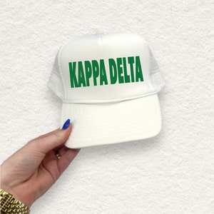 May include: A white trucker hat featuring the green text "KAPPA DELTA". The hat has a white mesh back and a curved peak. The hat is held by a person.