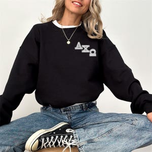 Op de afbeelding: Zwart sweatshirt met witte bies en de Griekse letters ΑΧΩ in zilver. Het sweatshirt wordt gedragen met een blauwe spijkerbroek en zwarte sneakers.