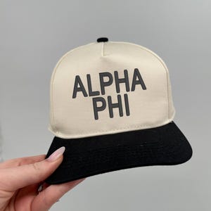 May include: A baseball cap with a cream-coloured crown and a black brim. The text "ALPHA PHI" is printed in black across the front of the cap. The cap has a black button on top.