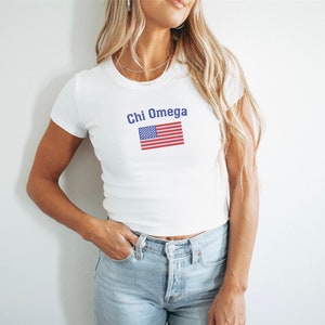 May include: White t-shirt with the text "Chi Omega" above an American flag graphic. The flag features red and white stripes and a blue square with white stars. The shirt is a fitted style.