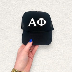May include: Black trucker hat with white Greek letters A and Phi on the front. The hat has a mesh back and a curved brim. The hat is held by a person.