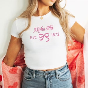 Puede incluir: Top corto blanco con el texto "Alpha Phi Est. 1872" en rosa y un lazo. Un cárdigan rosa con estampado de corazones rojos se lleva sobre los hombros.