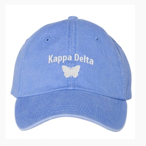 May include: A pale blue baseball cap featuring the embroidered text "Kappa Delta" and a white butterfly emblem. The cap has a curved peak and a six-panel design. The hat is made of a soft, washed cotton material.