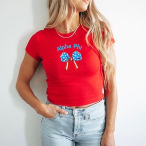 Puede incluir: Camiseta roja con un lazo azul y blanco y el texto "Alpha Phi". El diseño del lazo incorpora estrellas y rayas. La camiseta es de estilo ajustado.