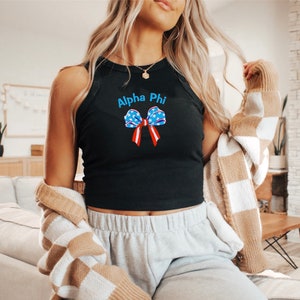 Puede incluir: Camiseta sin mangas negra con el texto "Alpha Phi" en azul y un lazo con diseño de estrellas y rayas. La camiseta sin mangas es sin mangas y tiene un cuello redondo.
