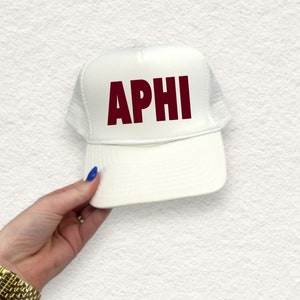 May include: White trucker hat with the letters "APHI" in maroon across the front. The hat has a mesh back and a curved brim. The hat is being held by a person with a gold watch and blue nail polish.