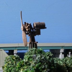 May include: A wooden model of a vintage propeller engine with a brown wooden propeller and a metal base. The engine is mounted on a wooden post and is surrounded by green foliage.