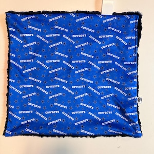May include: A blue and black baby blanket with the word "COWBOYS" in white, repeated across the surface with star accents. The blanket has a black, fluffy border and a white satin ribbon attached to the top.