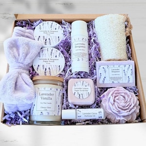 May include: A gift box containing a lavender-scented soy candle, body cream, sugar scrub, solid body butter, goat milk soap, sugar scrub bar, perfume oil, a loofah sponge, and a plush light purple headband, all nestled in purple paper shred.