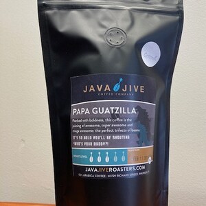 May include: A black coffee bag with the text "JAVA JIVE COFFEE COMPANY" and "PAPA GUATZILLA". The bag has a small round sticker that says "Ground". The bag is sitting on a wooden surface.